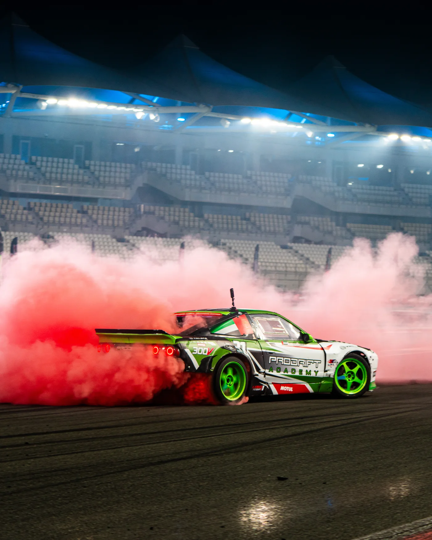 A Prodrift Academy car mid-drift trailing pink tyre smoke under stadium lights